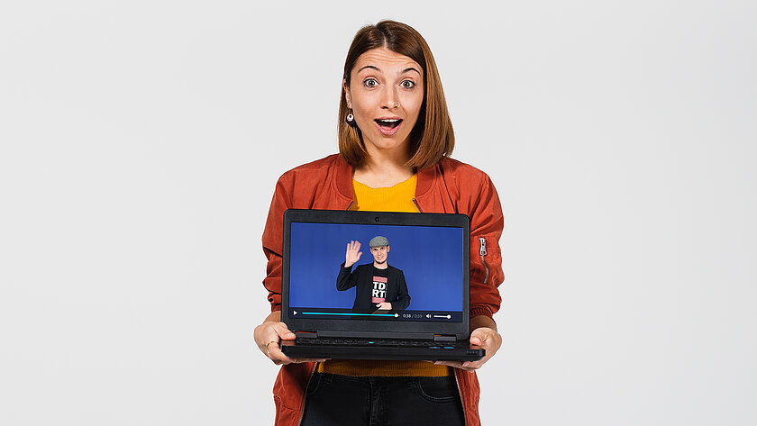 Frau mit begeistertem Gesichtsausdruck hält Laptop, auf dem das Begrüßungsvideo von Studienberater Thomas Bartl zu sehen ist. (öffnet Vergrößerung des Bildes)