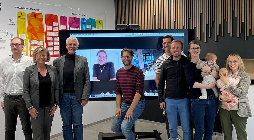 Das Bild zeigt eine Gruppe von Menschen, die vor einem Bildschirm stehen. Alle Personen lächeln. 
