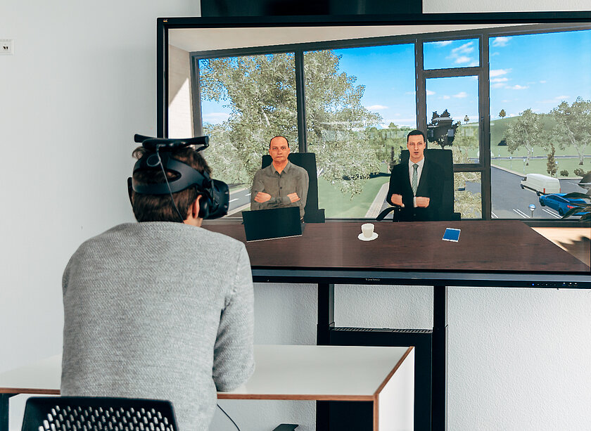 Das Bild zeigt eine Person mit einer virtuellen Brille an einem Schreibtisch vor einem Bildschirm sitzend. Auf dem Bildschirm sind zwei Personen zu erkennen, die virtuell der Person vor dem Bildschirm gegenüber sitzen. 